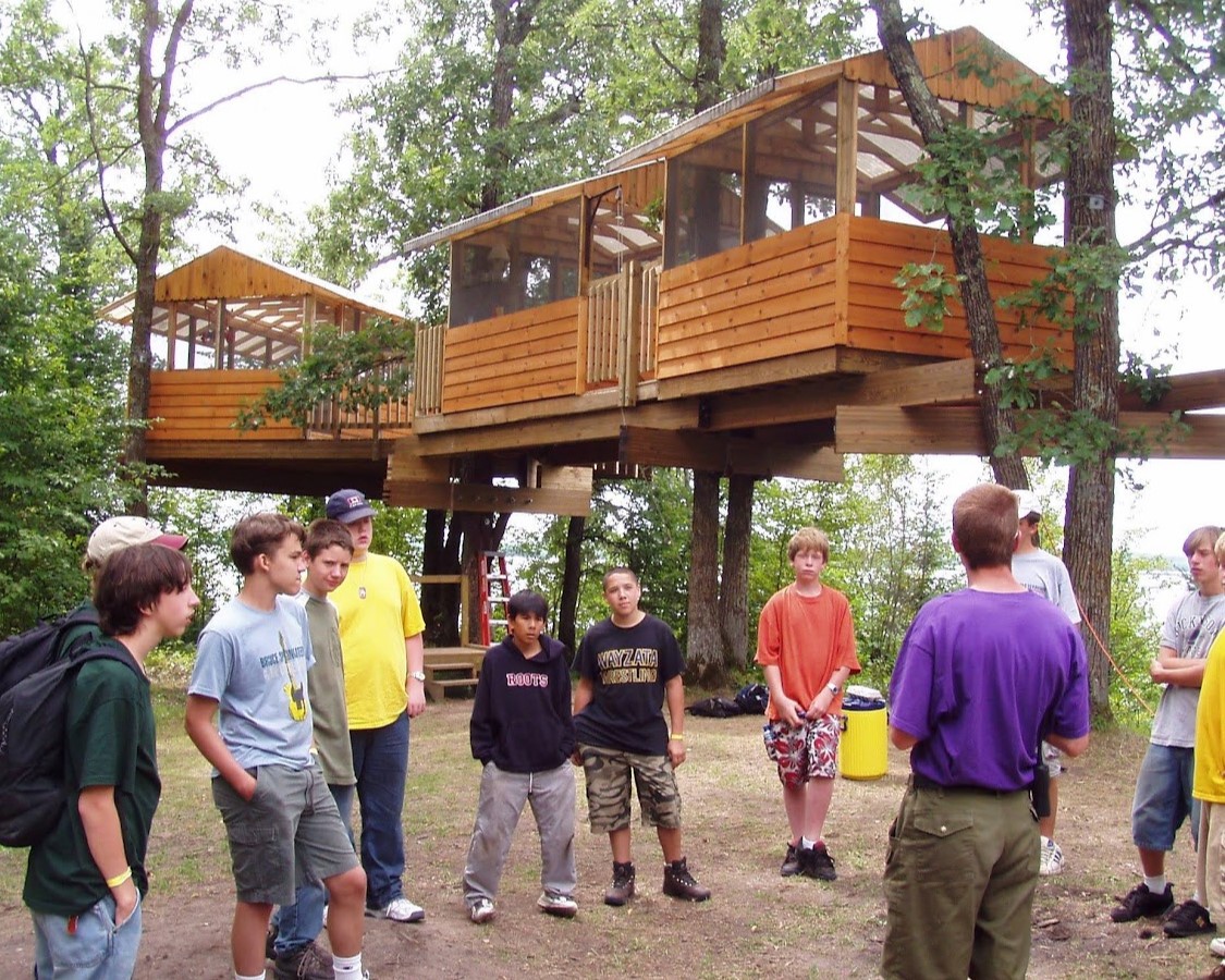 A group of Scouts are looking at the staff member who is leading some sort of talk. The treehouses are in the background, each one the size of a small house or very large RV