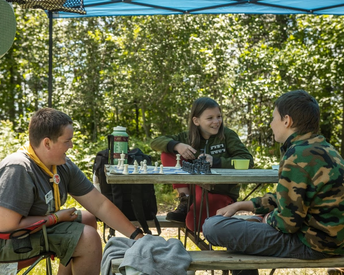 Three Scouts playing chess at a table in the shade