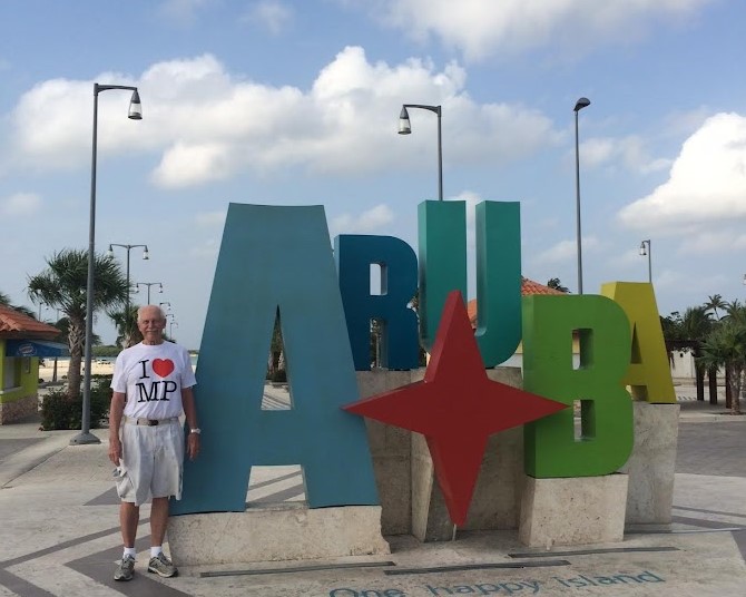 An older gentleman wearing an 'I heart Many Point' shirt in front of a sign that says 'Aruba'