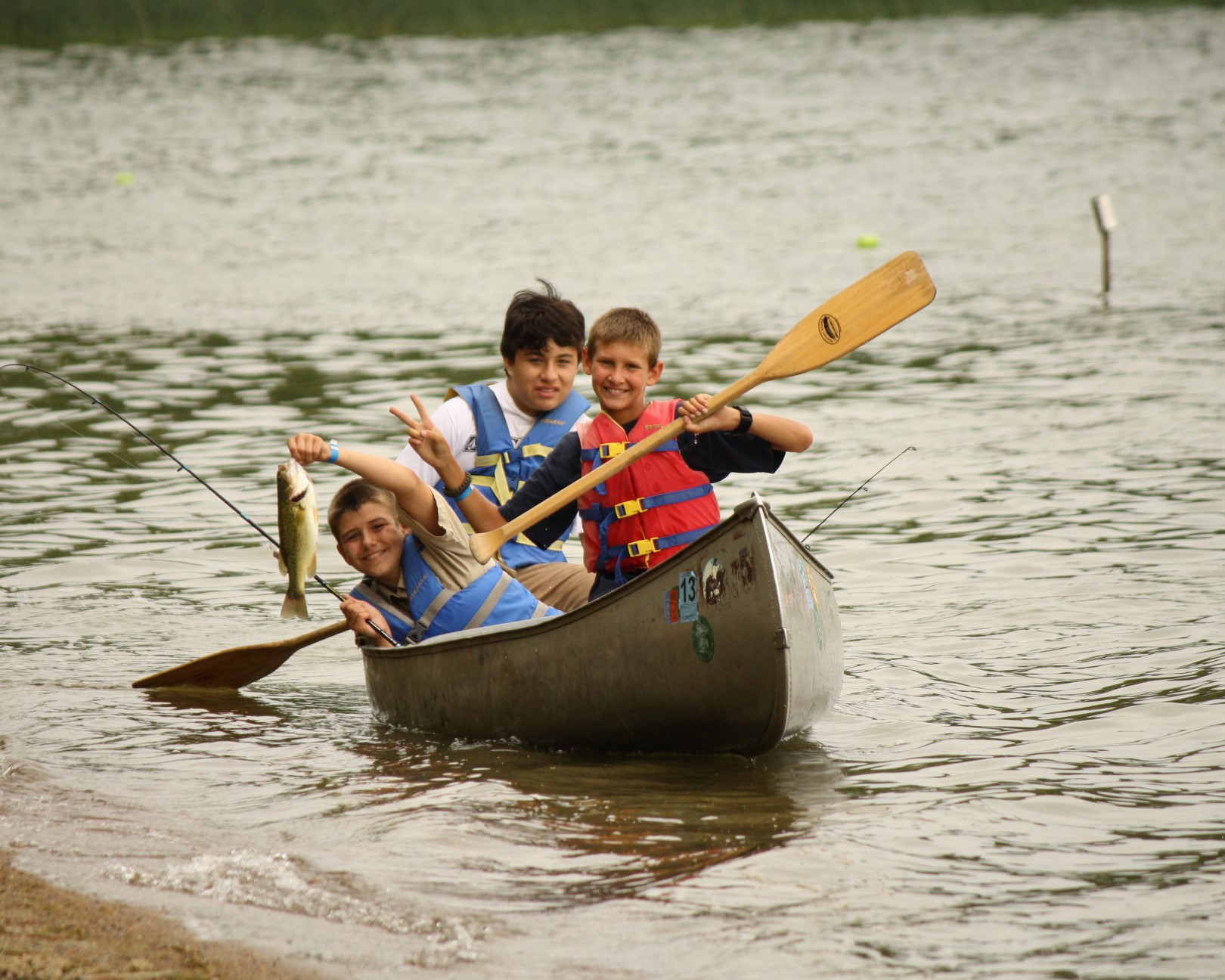 Three Scouts are in a canoe on the lake. One is holding a paddle, one is holding a fishing rod, and the third is holding a head-sized fish