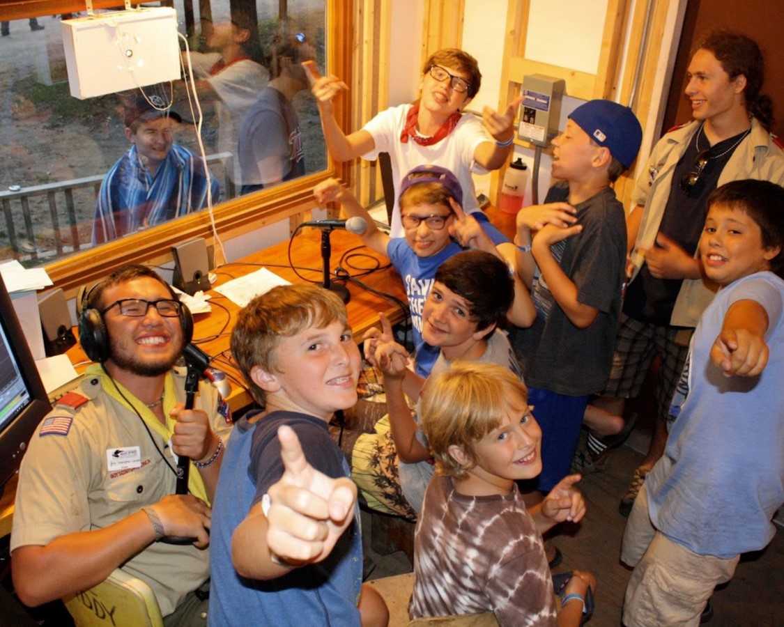 A group of Scouts smiling at and pointing to the camera while huddled around a desk with a microphone and script on it